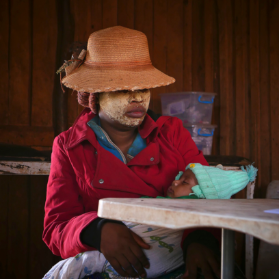 photo artistique d'une femme habillé d'une veste rouge et d'un chapeau sur la tête, son visage est enduit d'argile blanche pour protéger sa peau d soleil, elle est assise et porte dans ses bras son bébé qui dort - Agrandir l'image, fenêtre modale